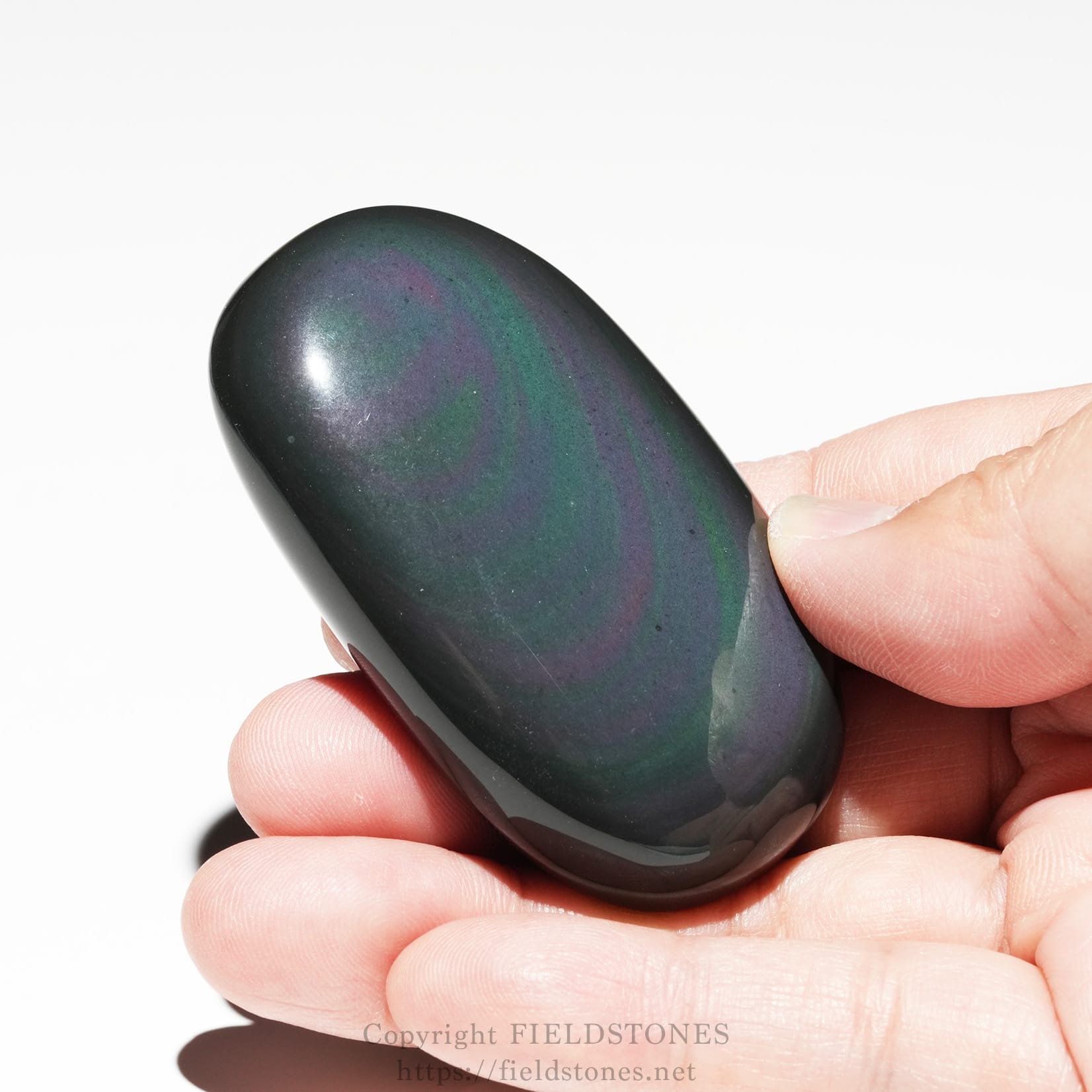 6682-4 - FieldStones Close-up of a smooth, oval palm stone with purple and green swirls being held between a thumb and finger