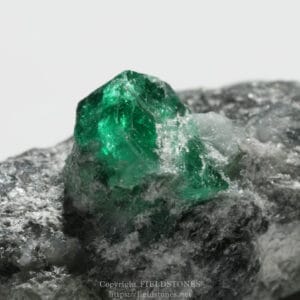 Close-up of a bright green emerald crystal embedded in a gray rocky matrix, sparkling facets.