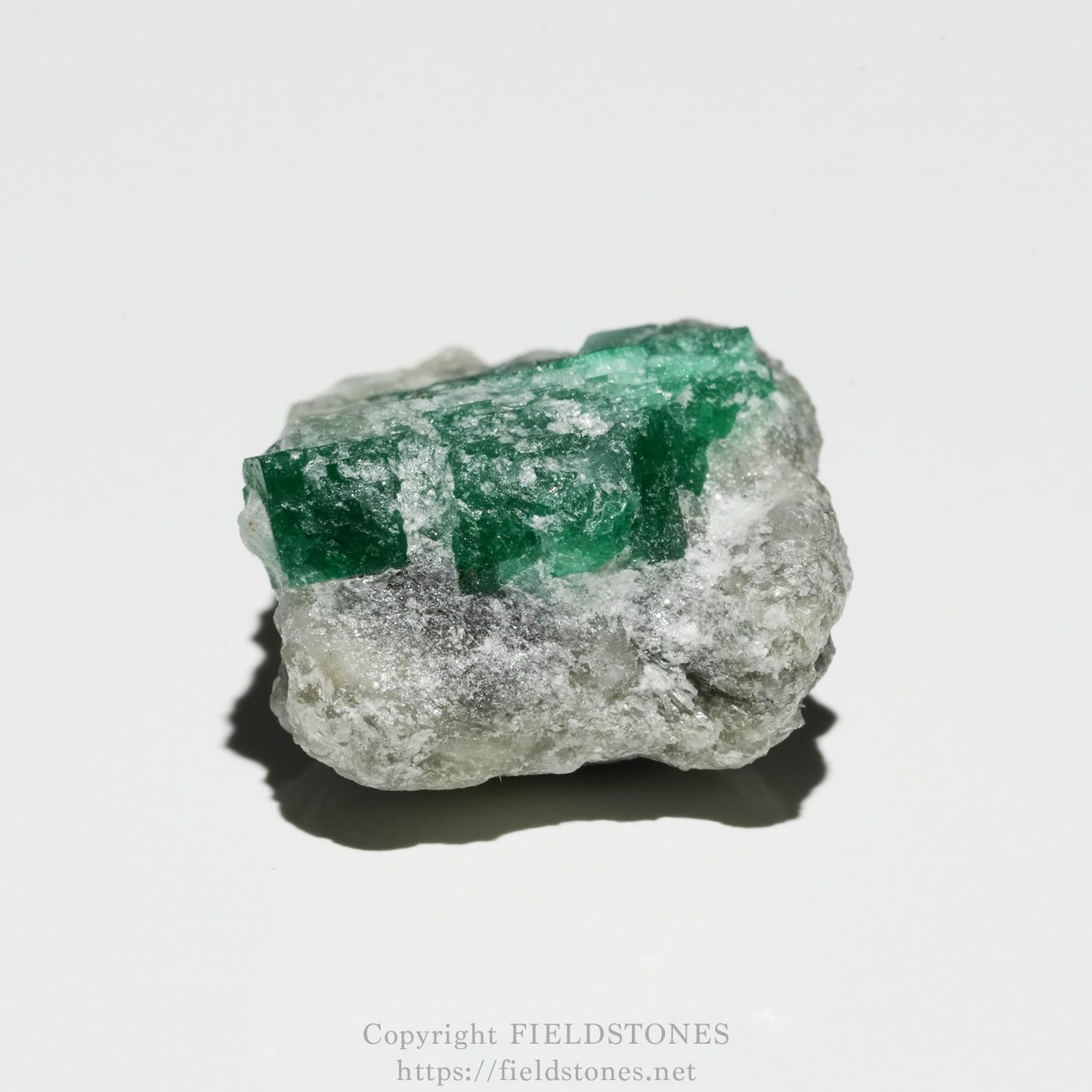 6700-2 - FieldStones Rough green emerald crystal on a gray rock matrix, set against a white background.