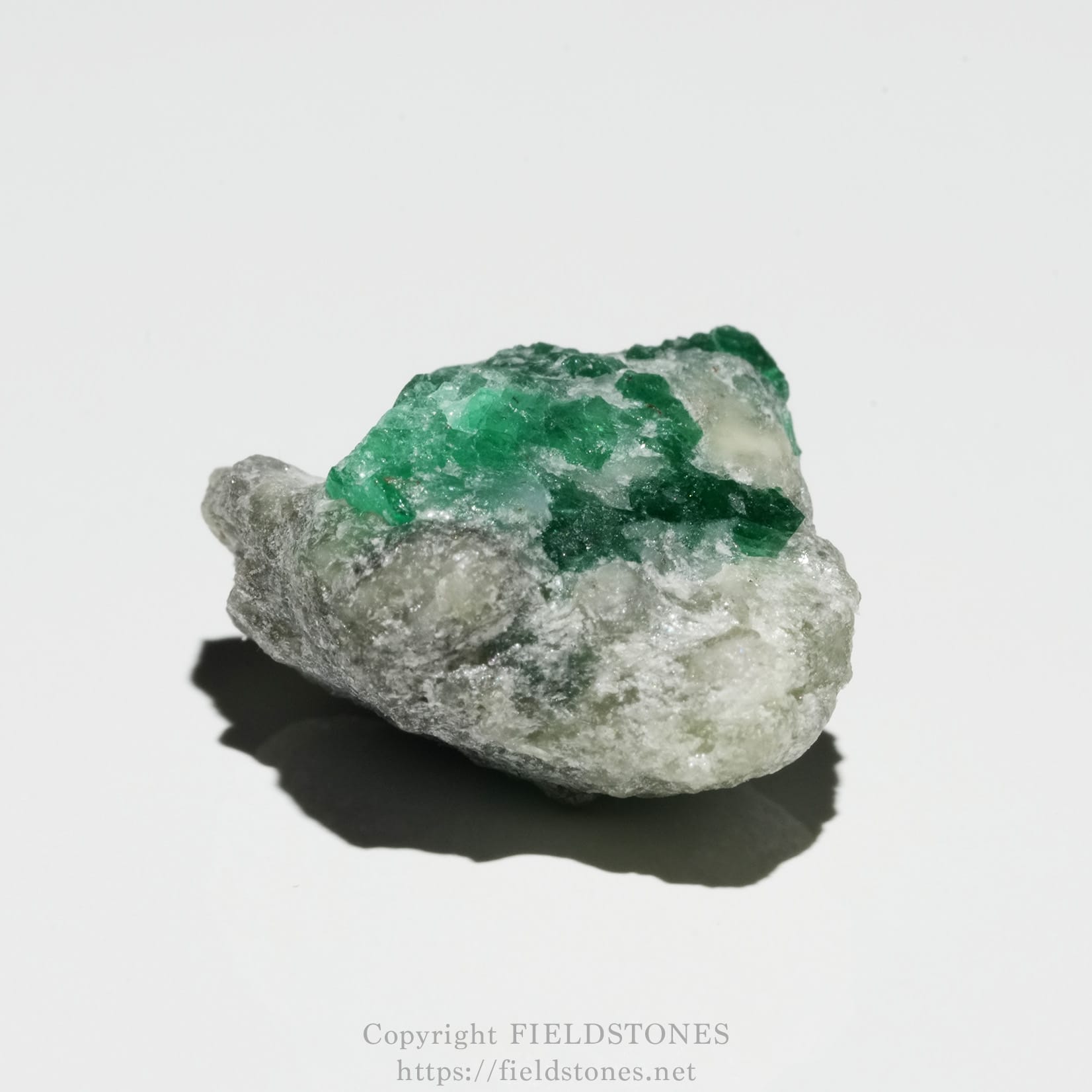 6700-5 - FieldStones Rough gray rock with bright green emerald crystals clustered on top against a white background.