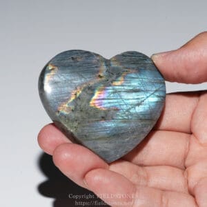 Heart-shaped labradorite stone held between fingers, displaying a blue iridescent rainbow sheen.