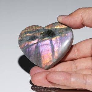 Close-up of a polished, heart-shaped labradorite stone showing iridescent pink, purple, and blue hues being held between two fingers by a light-gray background.
