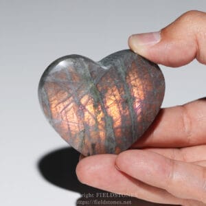 Heart-shaped labradorite stone held between a thumb and finger, showing iridescent orange-blue flashes.