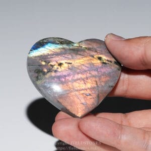Heart-shaped, polished paua shell displaying blue, green, and pink iridescence being held between two fingers.