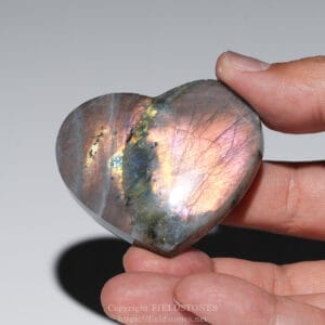 Polished heart-shaped labradorite stone held between a thumb and finger, showing iridescent rainbow colors for the image's meaning as a gemstone:**
