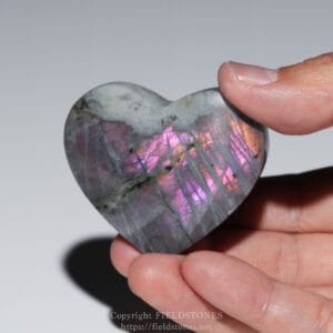 Hand holding a polished heart-shaped labradorite stone with iridescent pink and purple sheen.