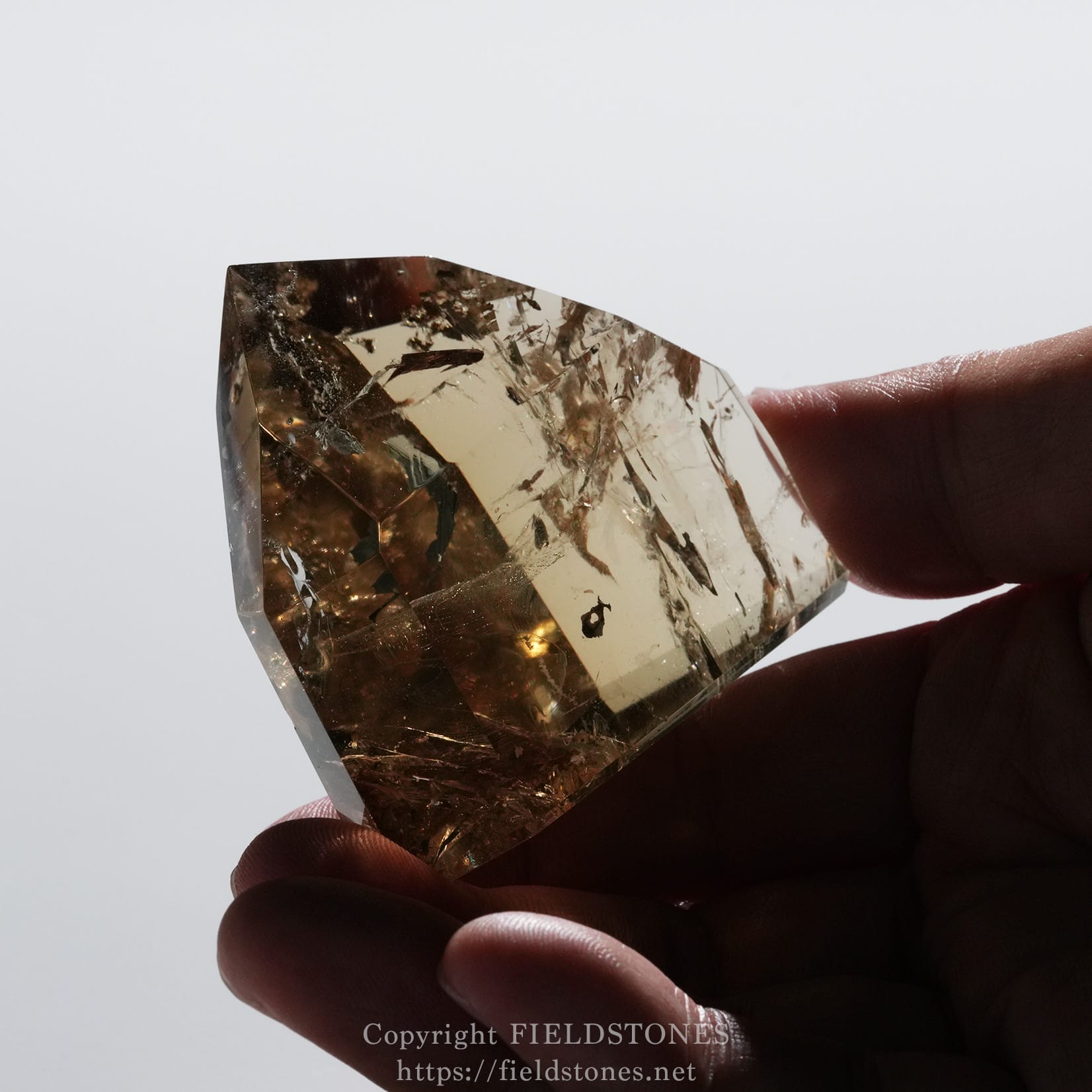 6708-11 - FieldStones Close-up of an amber-colored faceted crystal held between fingertips against a pale background, showing light through the facets.