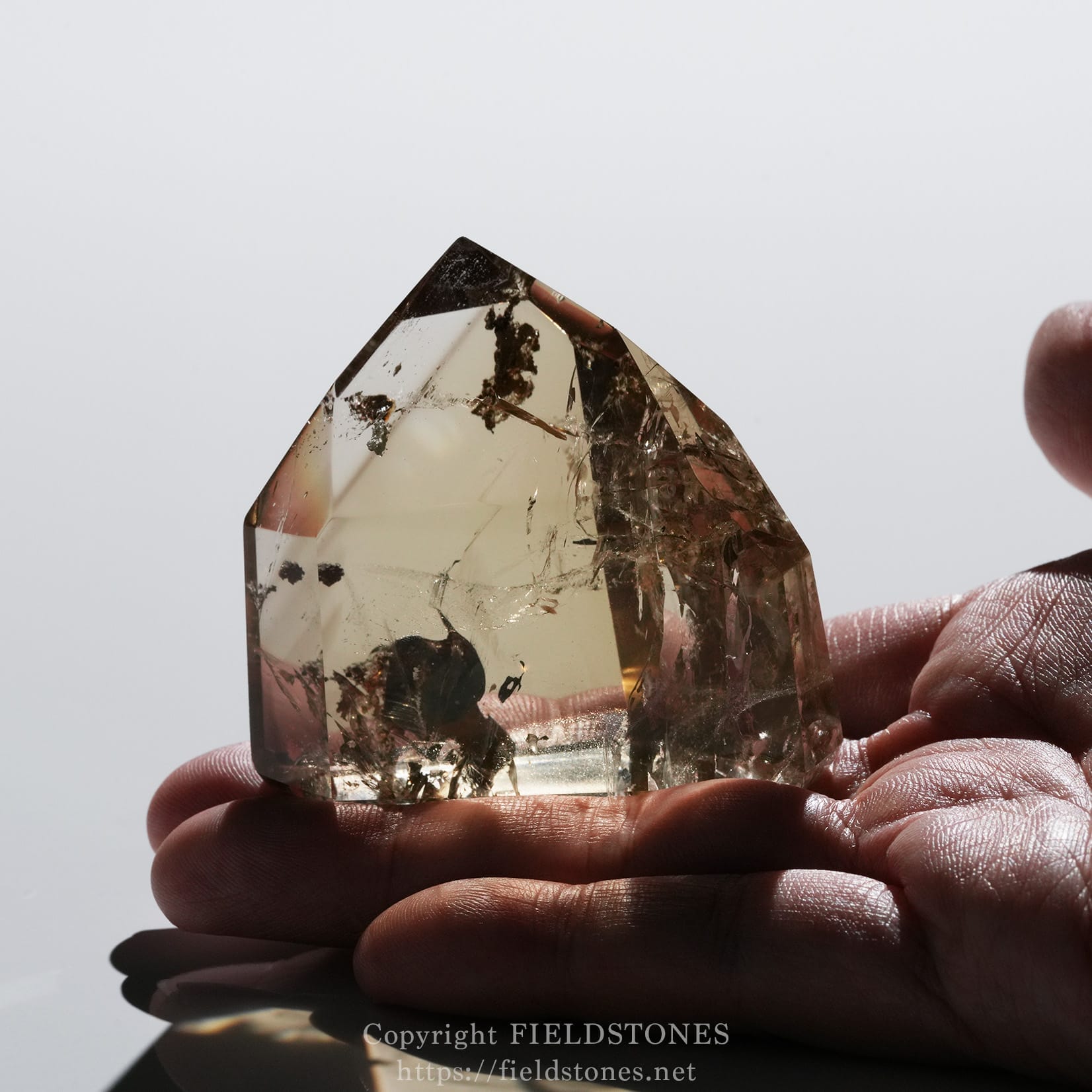 6708-12 - FieldStones Close-up of a clear quartz crystal held between fingers, showing internal fractures and dark inclusions against a reflective surface.