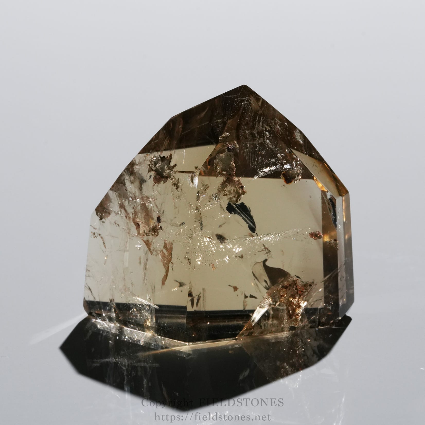 6708-4 - FieldStones Close-up of a polished brown crystal point with faceted faces and internal inclusions, resting on a black base against a light gray background.