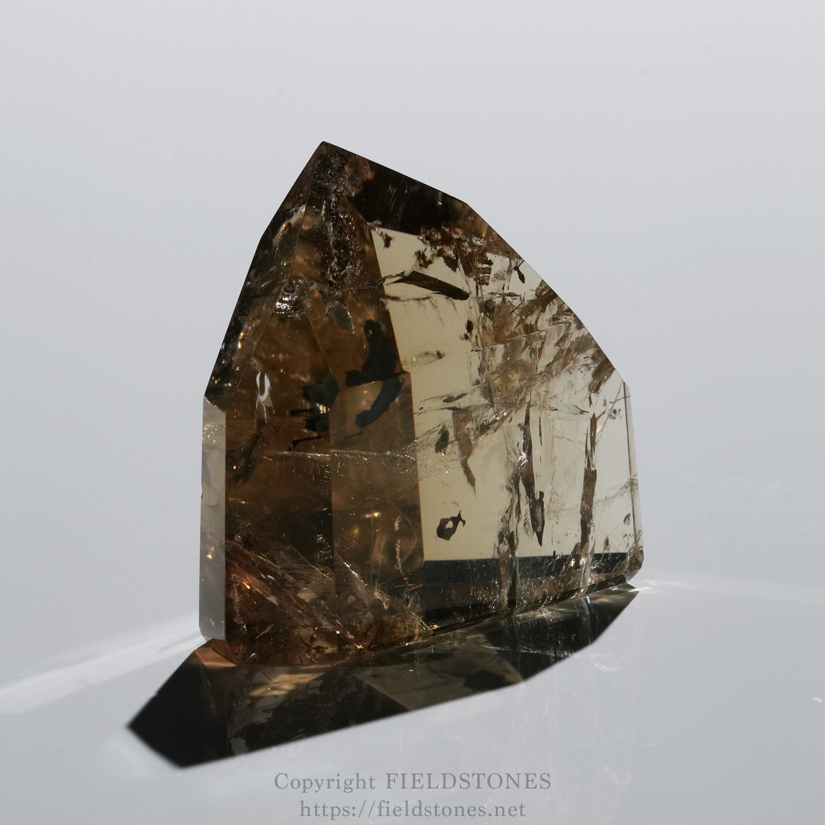 6708-6 - FieldStones Brown smoky quartz crystal standing upright on a reflective surface with faceted sides against a pale background.”,”A translucent brown crystal with a pointed tip and jagged facets on a glossy surface.”,”Close-up of a brown prism crystal showing angular facets and reflections.”,”Large brown gemstone crystal with a pointed apex mounted on a mirror-like base.”,”A single brown crystal specimen with sharp edges, set against a light backdrop.