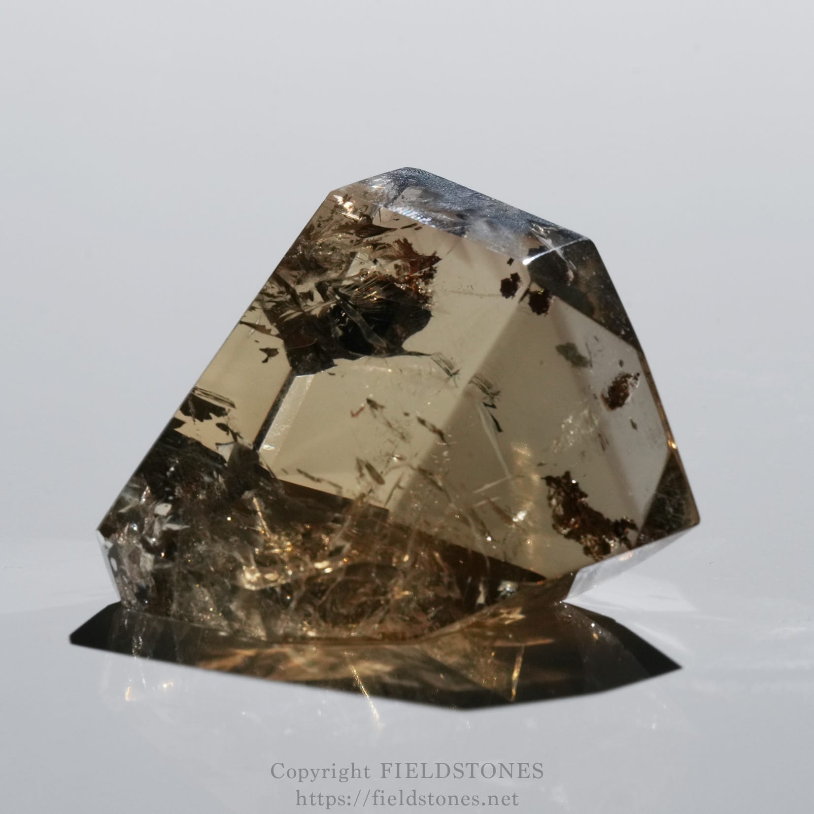6708-7 - FieldStones Faceted smoky quartz crystal on a white surface with sharp edges and brown inclusions.