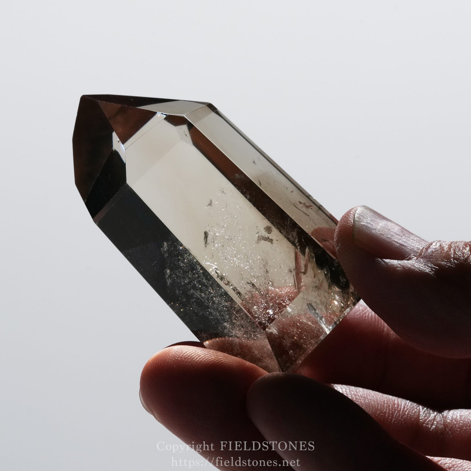 6709-11 - FieldStones Close-up of a clear, faceted crystal held between fingers against a pale background.