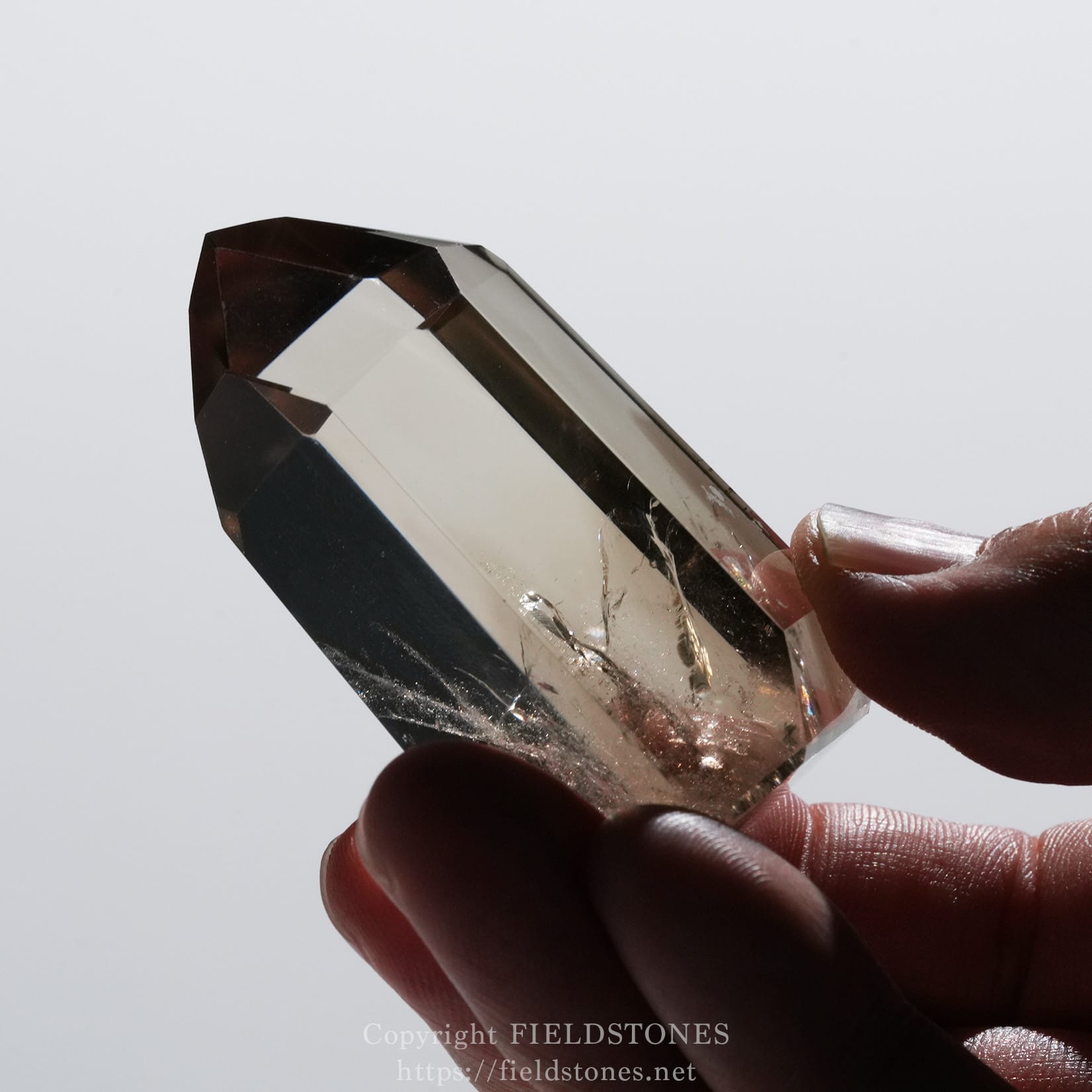 6709-12 - FieldStones Close-up of a faceted crystal prism held between fingers against a pale background, showing sharp angular facets and clarity.