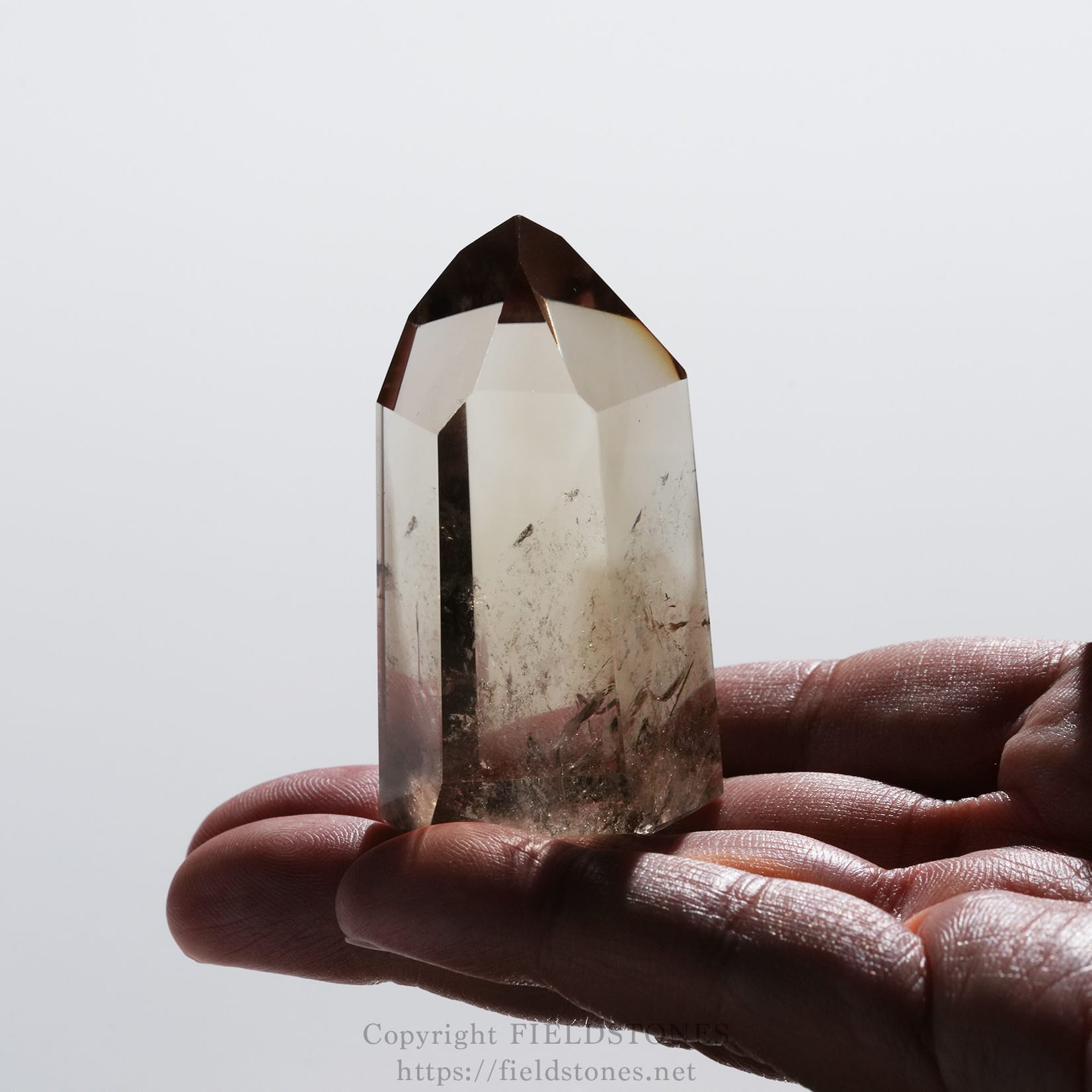 6709-13 - FieldStones Clear quartz crystal point held between fingers against a pale background, showing its facets and slight inclusions.