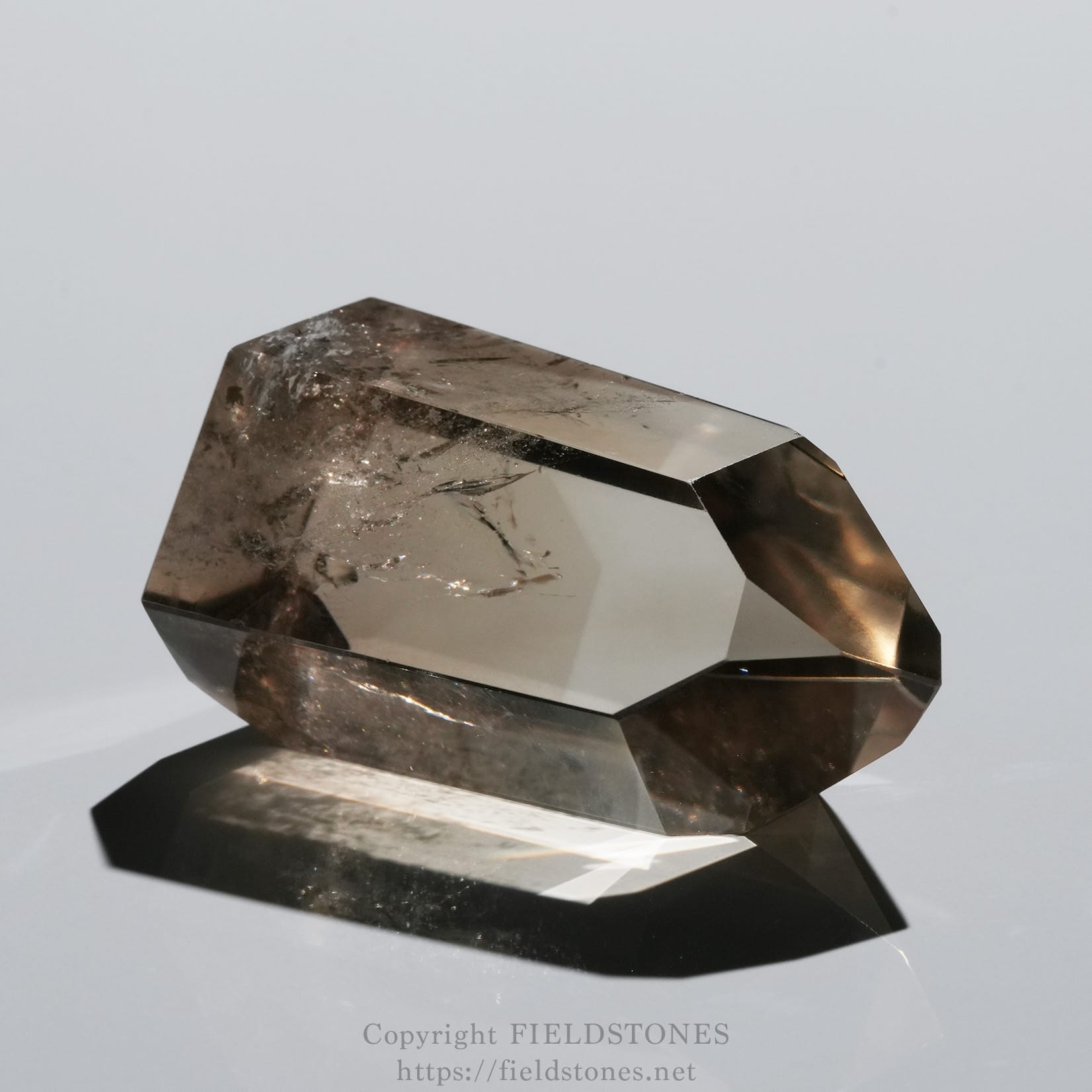 6709-2 - FieldStones Close-up of a faceted smoky quartz crystal on a reflective surface, showing sharp edges and subtle inclusions against a gray background.