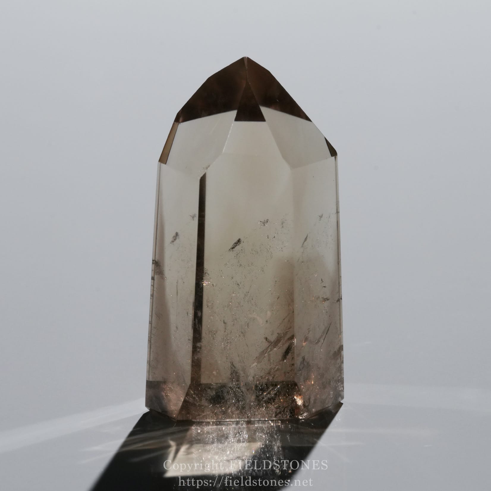 6709-7 - FieldStones Close-up of a tall clear quartz crystal with a pointed tip and a brown cap at the top, set against white background.