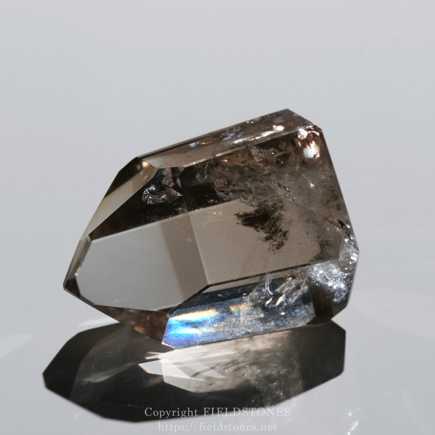 6710-1 - FieldStones Close-up of a faceted smoky quartz crystal on a dark stand, reflecting on a glossy surface.
