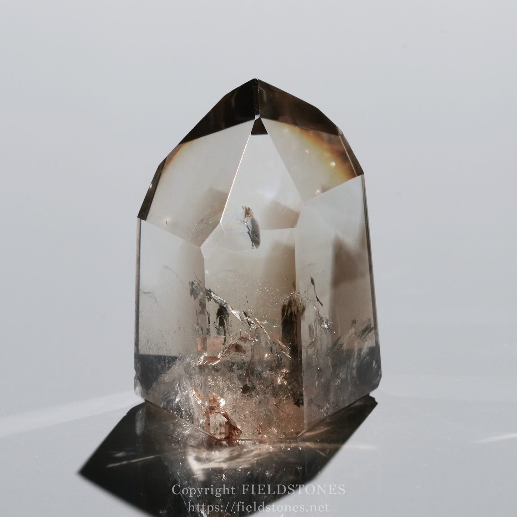 6710-4 - FieldStones Clear quartz crystal point with a rugged base, standing upright on a reflective surface, showing inner inclusions and subtle rainbow reflections.