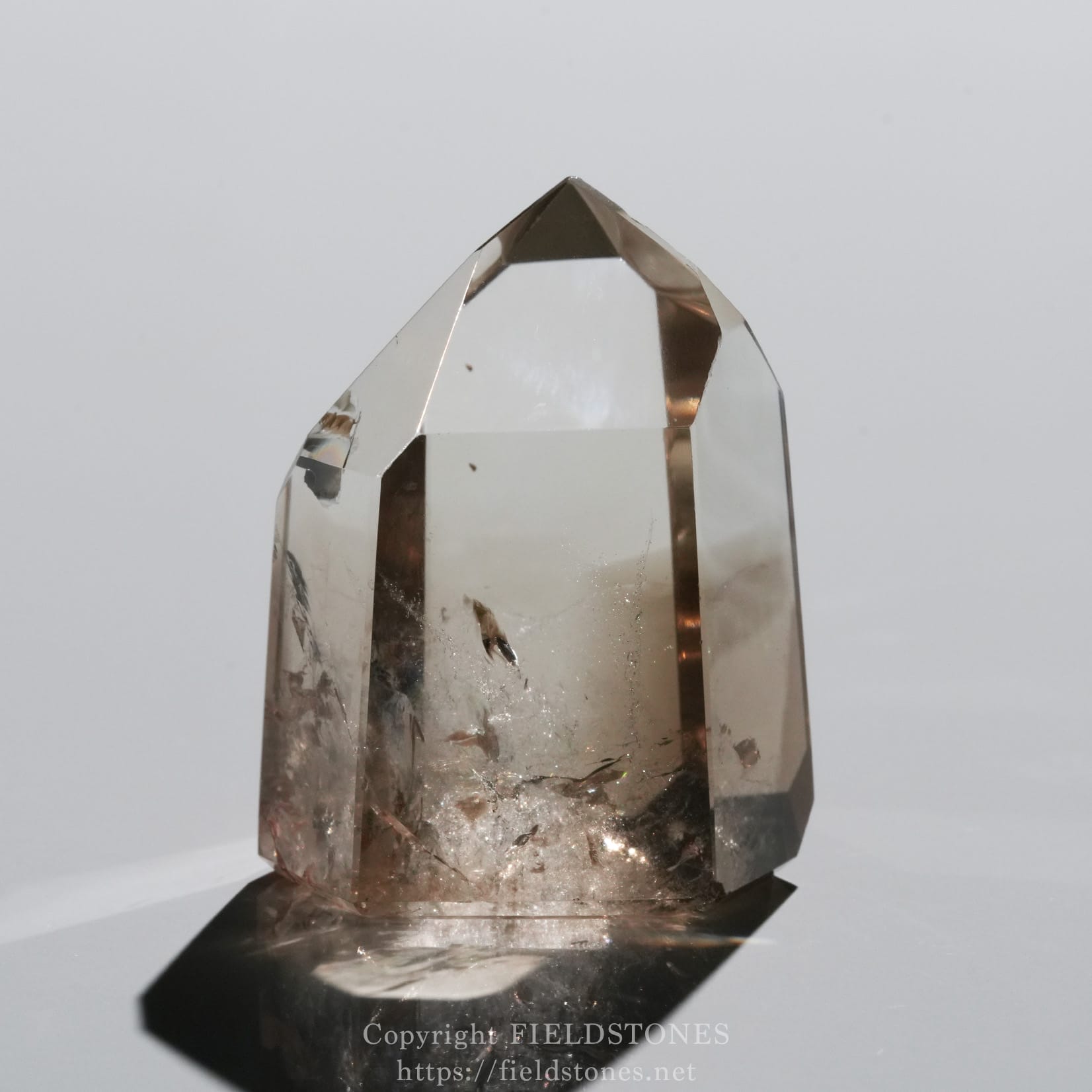 6710-5 - FieldStones Clear quartz crystal point perched on a dark stand, showing a faceted, pyramid-like tip against a pale background.