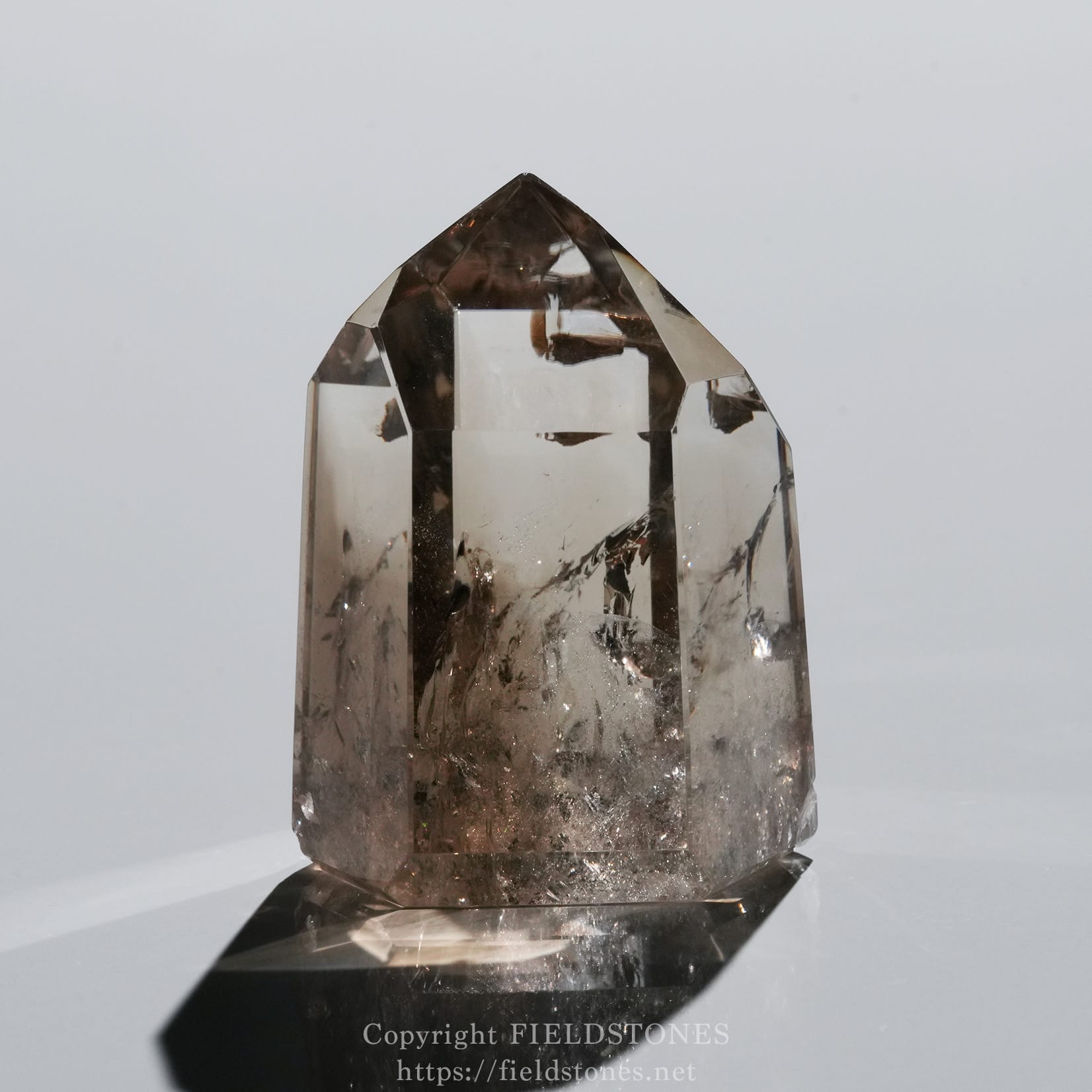 6710-7 - FieldStones Smoky quartz crystal point standing upright on a reflective surface, showing faceted edges and a cloudy interior.