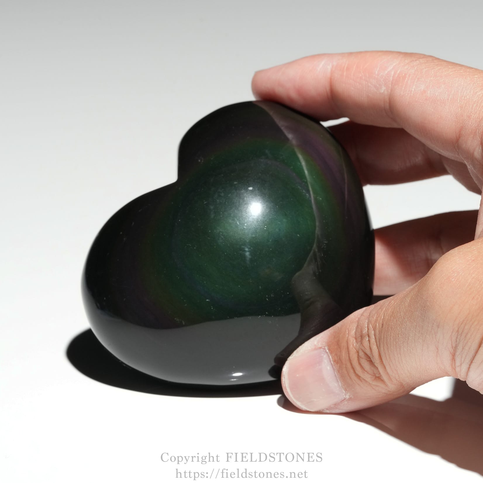 6711-6 - FieldStones Close-up of a dark green, heart-shaped polished stone held between the thumb and index finger against a light background