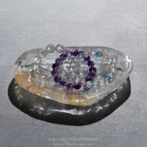 Beaded bracelets in purple, blue, and pink tones arranged inside a clear crystal shell on a gray surface.