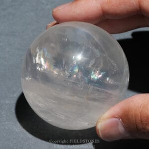 Close-up of a translucent quartz sphere held between a thumb and finger, showing subtle internal fractures and rainbow reflections inside.