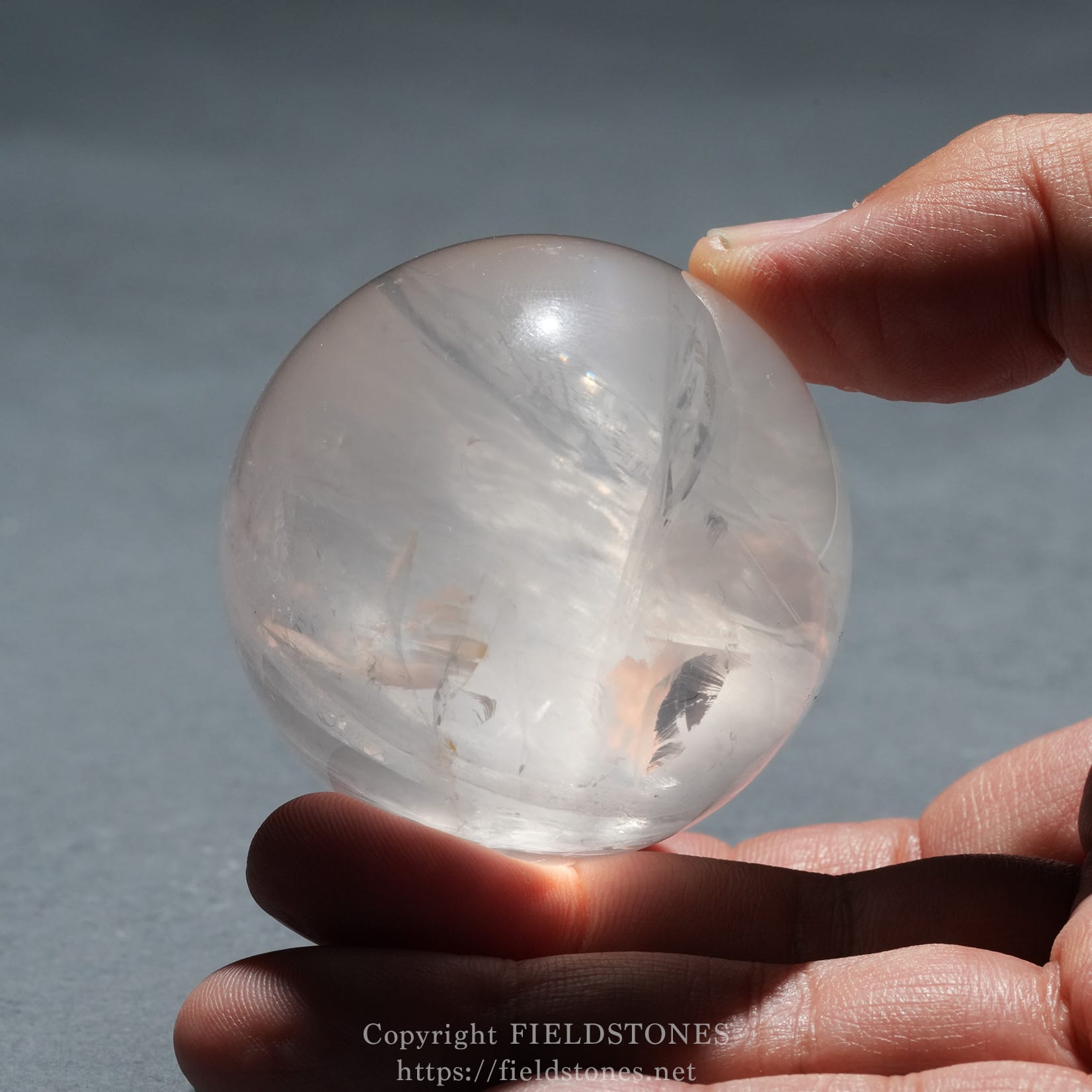 6719-10 - FieldStones Close-up of a hand pinching a clear glass sphere outdoors, with a blue-gray background.