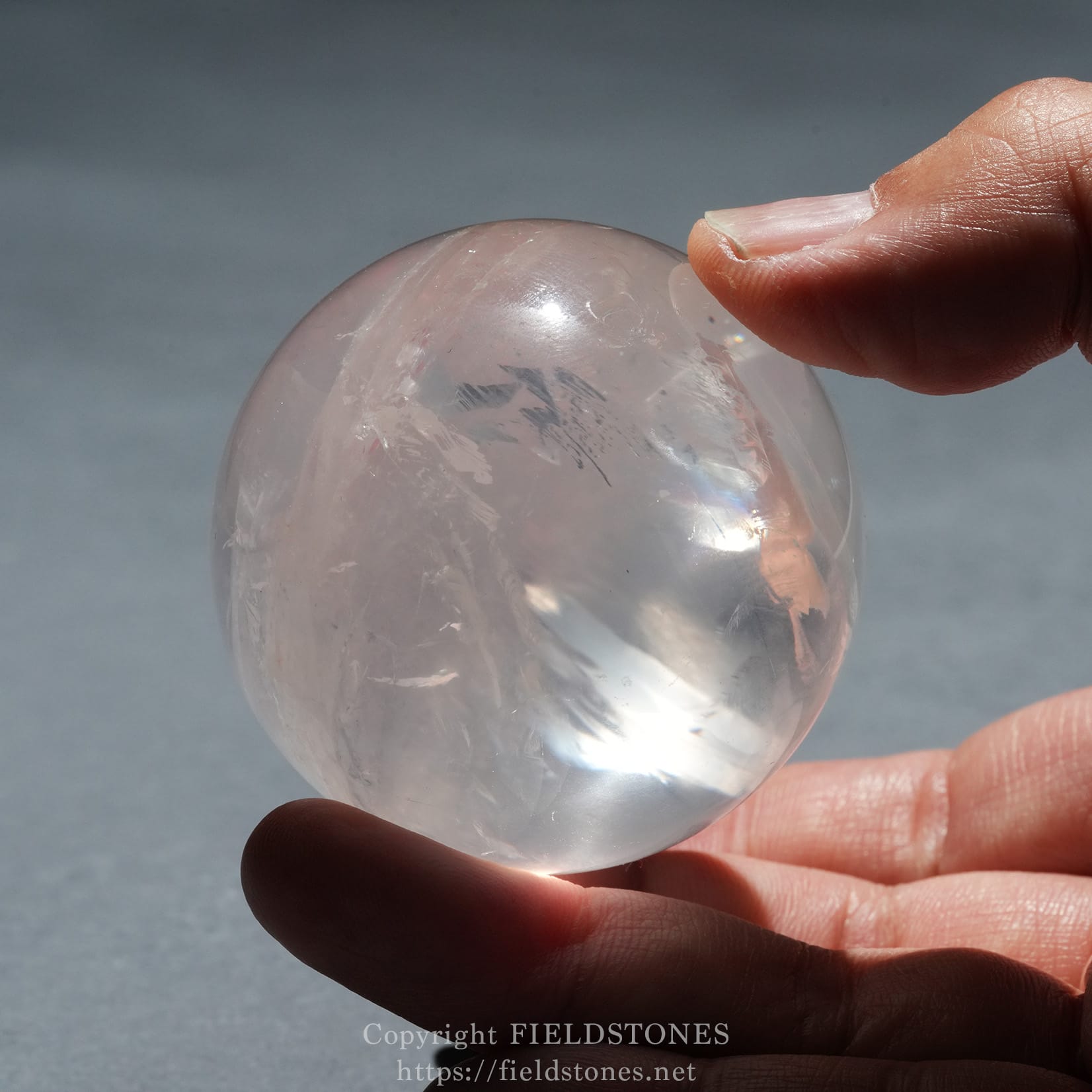 6719-11 - FieldStones Close-up of a clear crystal ball held between fingers against a soft blue background, with light reflections on the glass.