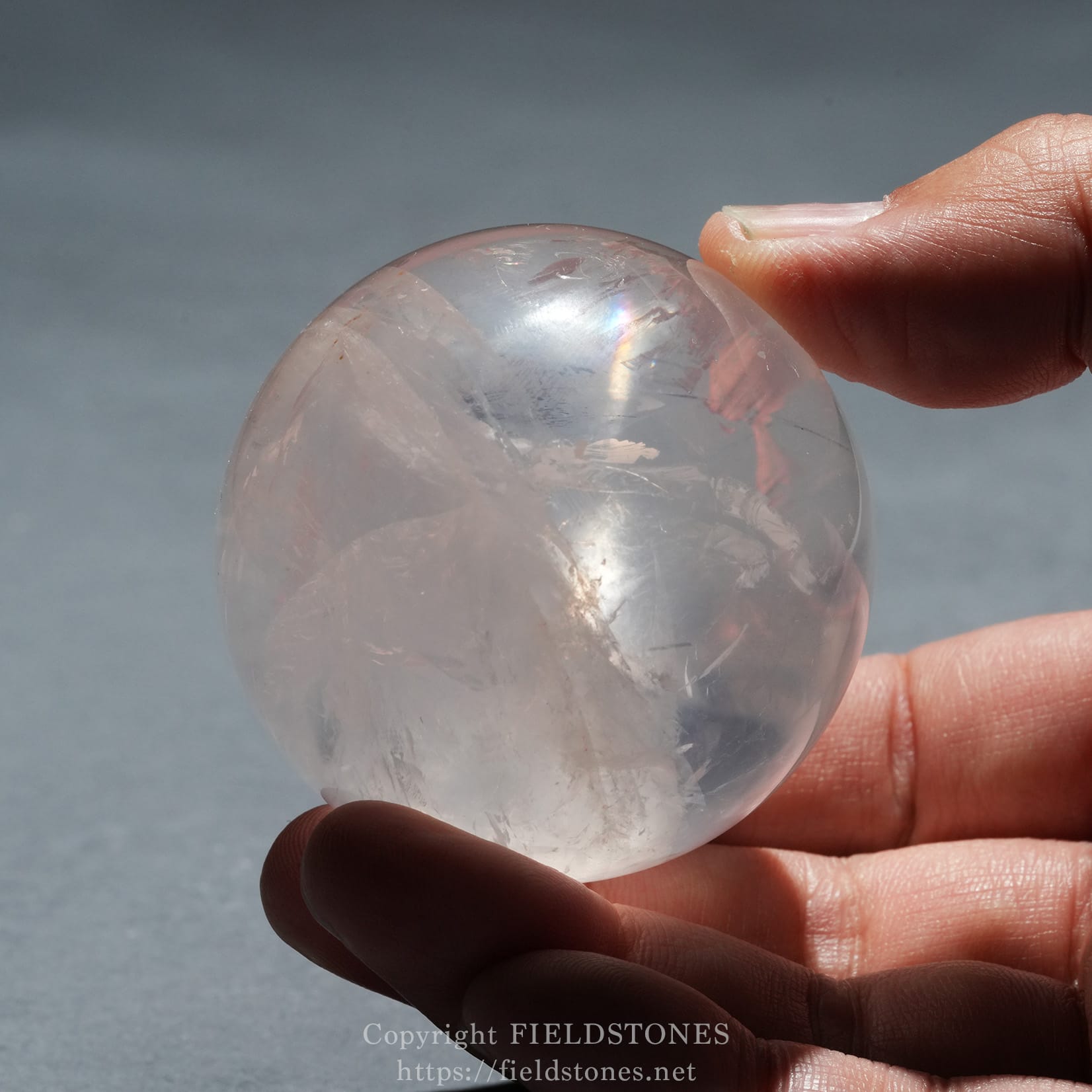 6719-12 - FieldStones Clear crystal ball held between fingers with light refractions, outdoors against a gray sky.