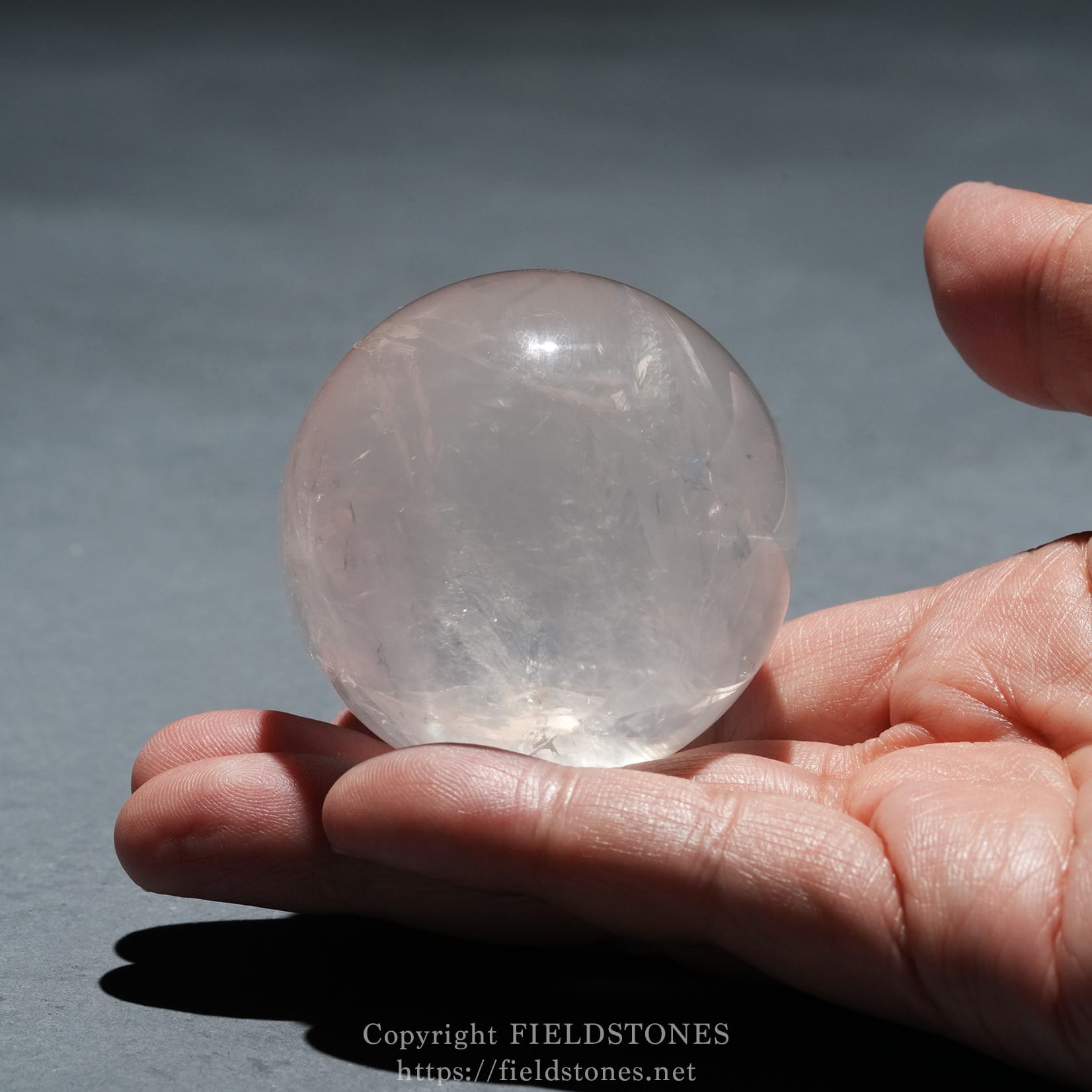 6719-13 - FieldStones Close-up of a clear crystal ball held between fingertips outdoors, with light reflections inside the sphere.