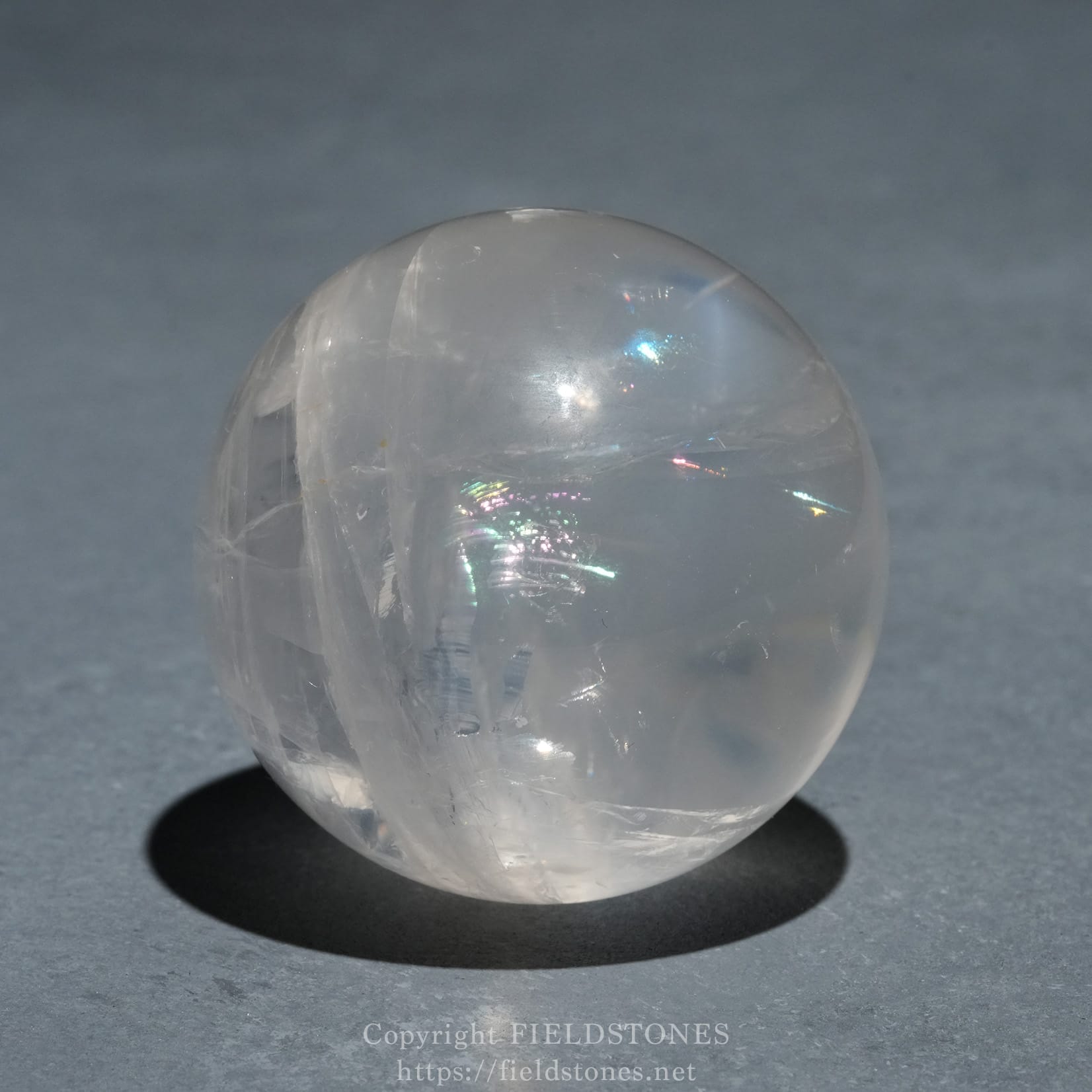 6719-2 - FieldStones Clear glass sphere resting on a dark gray surface, with a soft shadow underneath and subtle light reflections on the ball's surface.