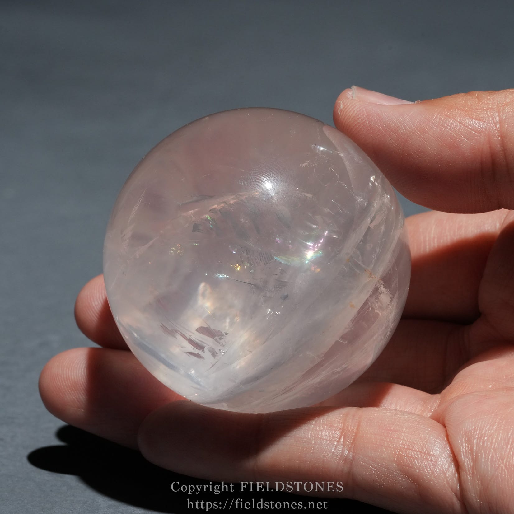 6719-4 - FieldStones Close-up of a translucent quartz crystal sphere held between two fingers with light reflecting inside the crystal against a gray background