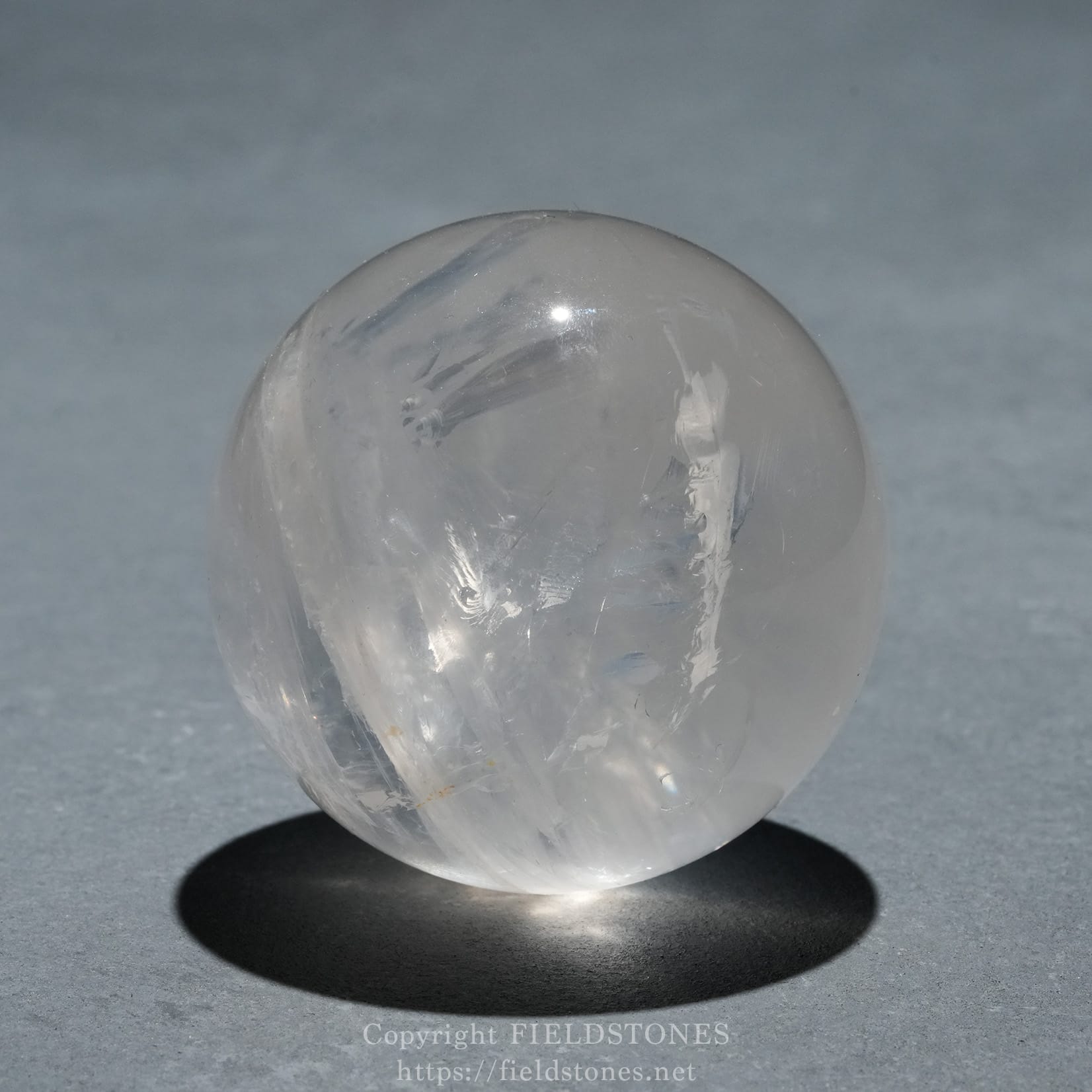 6719-5 - FieldStones Clear quartz crystal ball resting on a small stand, casting a dark shadow on a gray surface against a blue-gray background.