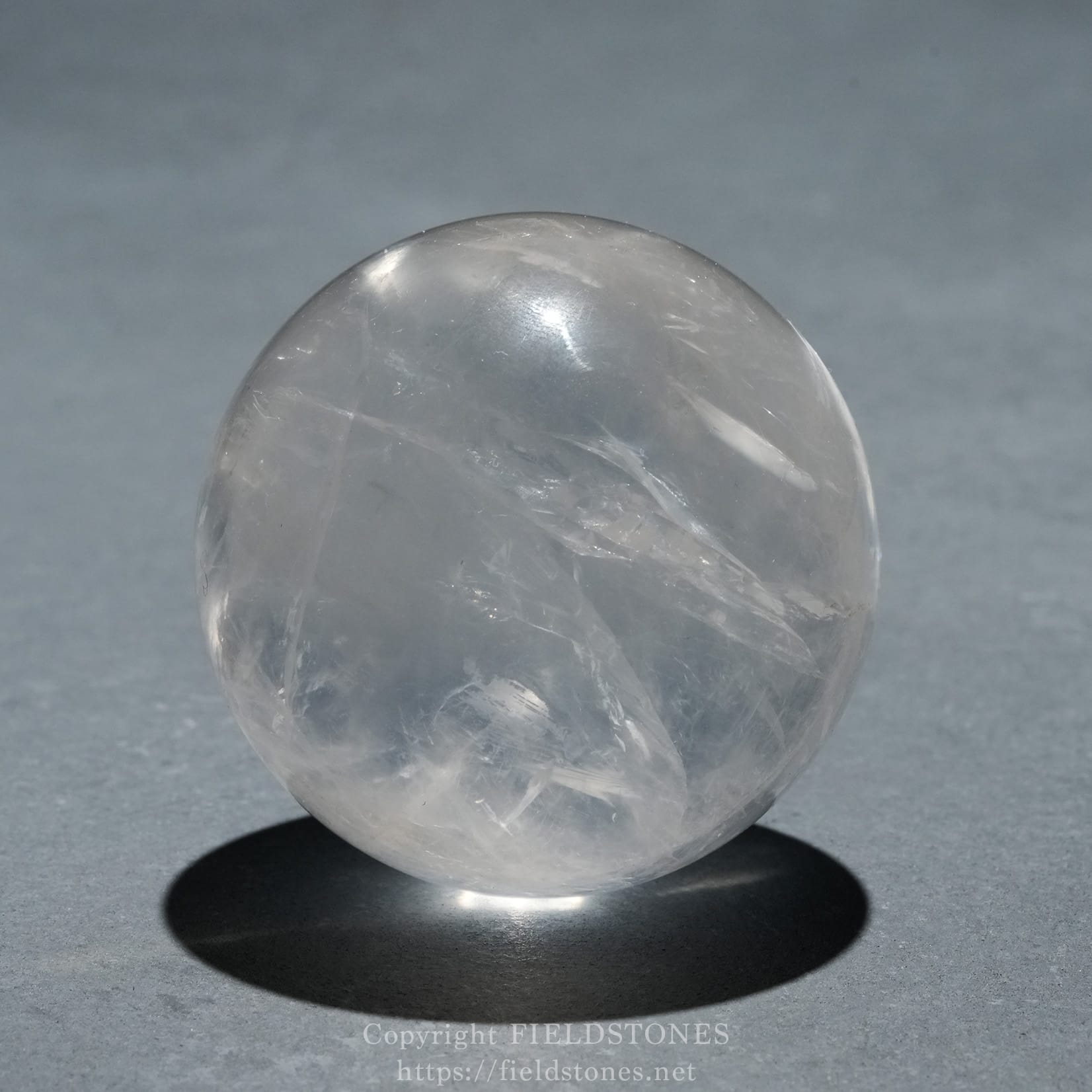 6719-6 - FieldStones Clear quartz crystal ball resting on a small black base, casting a circular shadow on a gray surface.