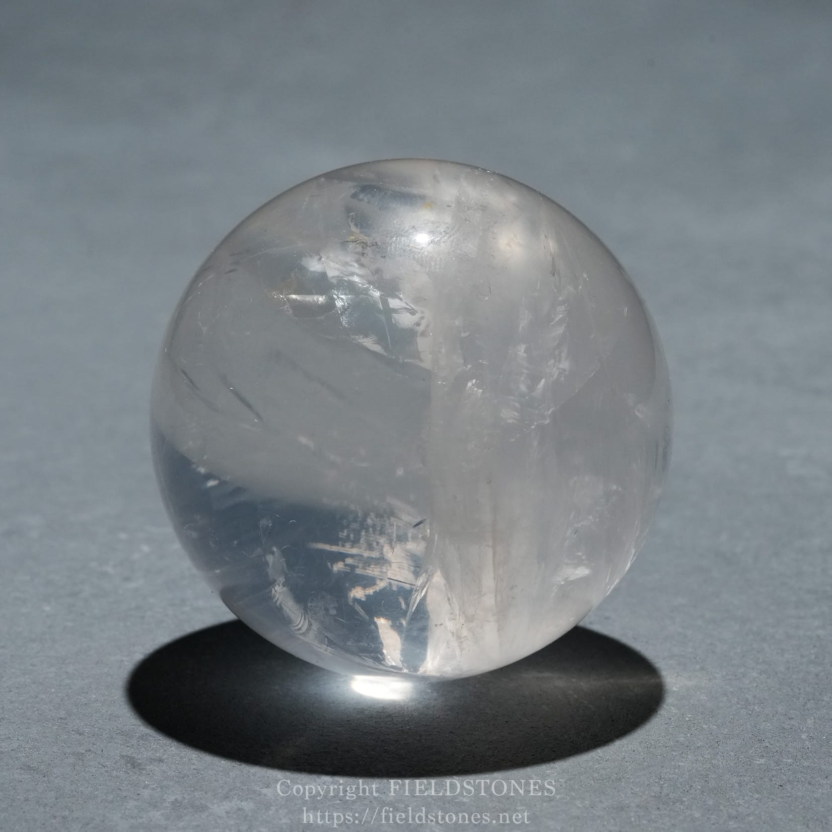 6719-7 - FieldStones Clear quartz sphere resting on a dark base with a soft shadow on a gray surface.