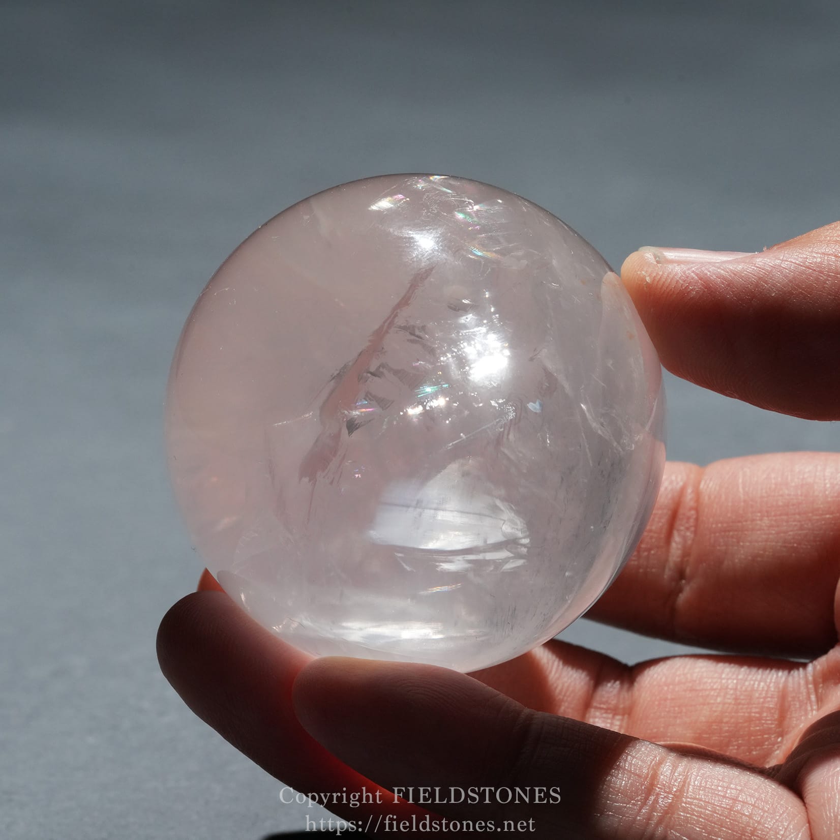 6719-9 - FieldStones Close-up of a pale pink quartz sphere being held between fingertips against a gray background, showing internal facets and reflections.