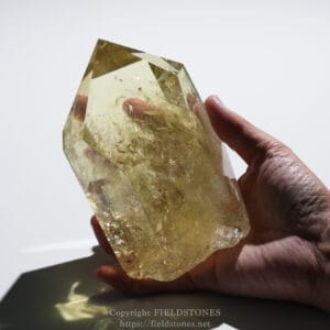 A hand holding a large clear quartz crystal with yellow tint and multiple faceted faces visible against a light background.