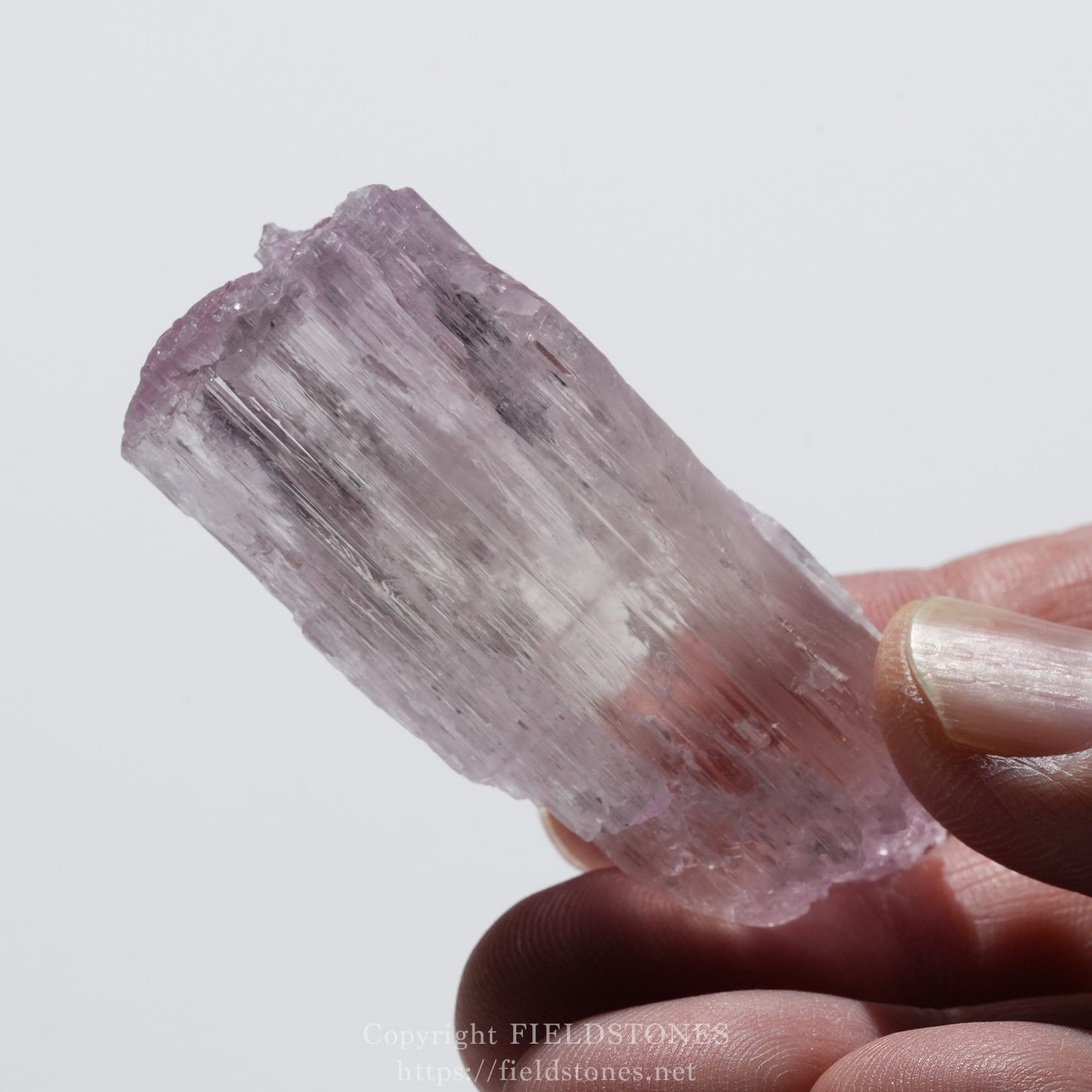 6726-8 - FieldStones Close-up of a rough pink-purple quartz crystal being held between a thumb and forefinger, showing its layered, striated surfaces.