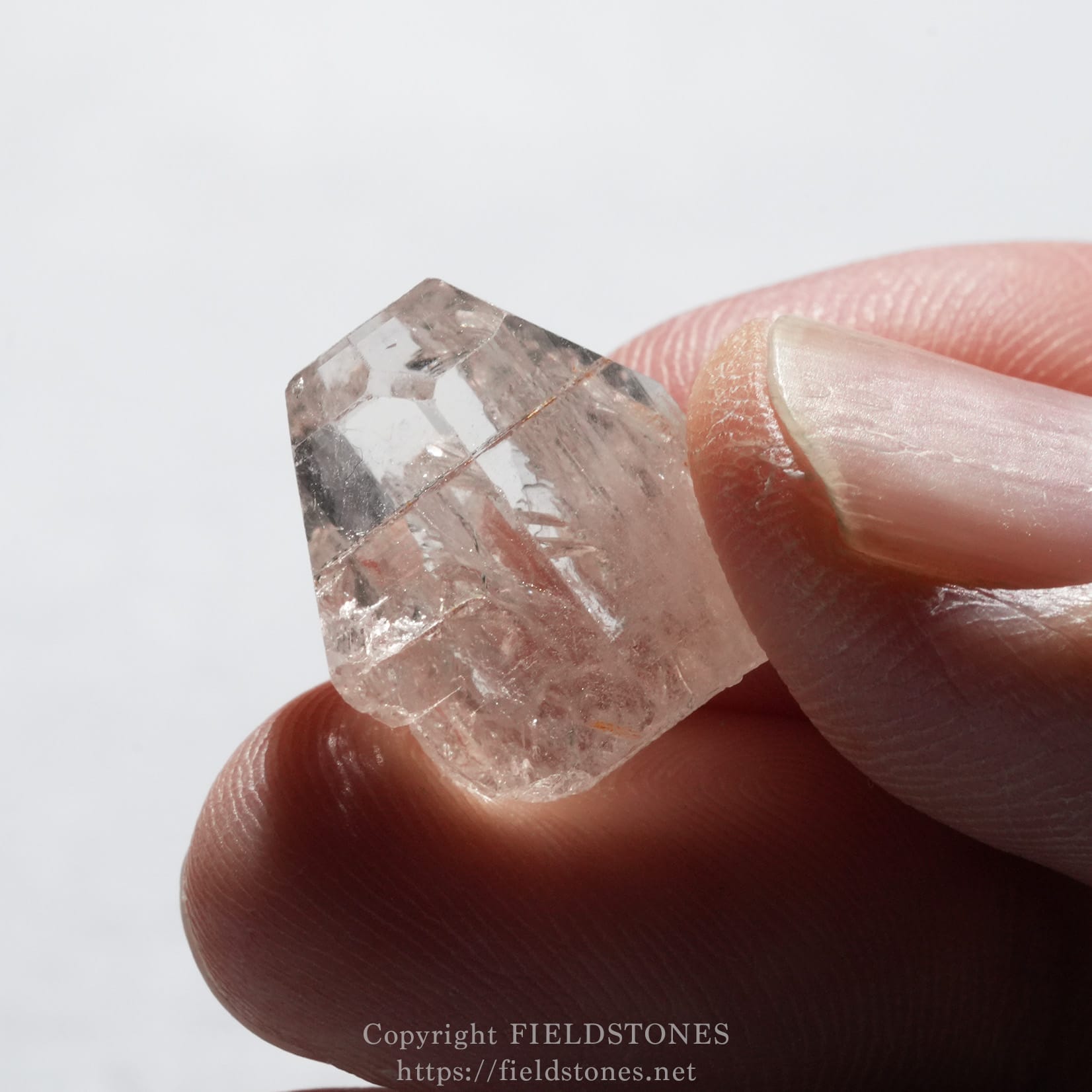 6737-8 - FieldStones Close-up of a pink rose quartz crystal being held between a thumb and forefinger, showing faceted edges and translucent facets.
