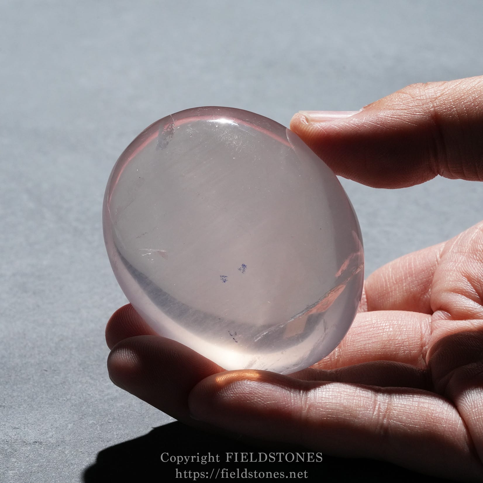 6740-3 - FieldStones Close-up of a hand holding a smooth pink quartz sphere outdoors against a pale surface, showing a glossy, translucent finish.