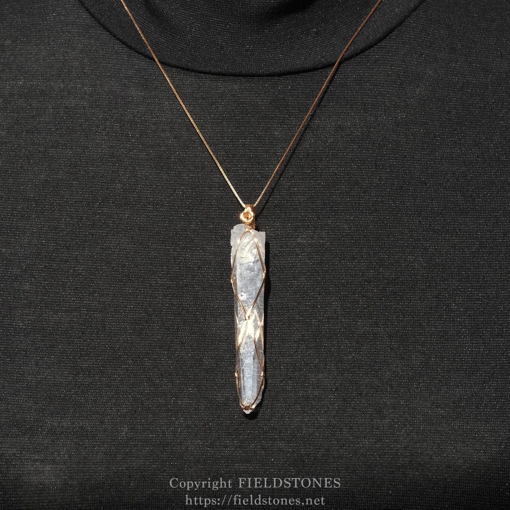 6751-10 - FieldStones Gold chain necklace with a copper-wrapped crystal pendant resting on black fabric; a vertical quartz point suspended by wire.