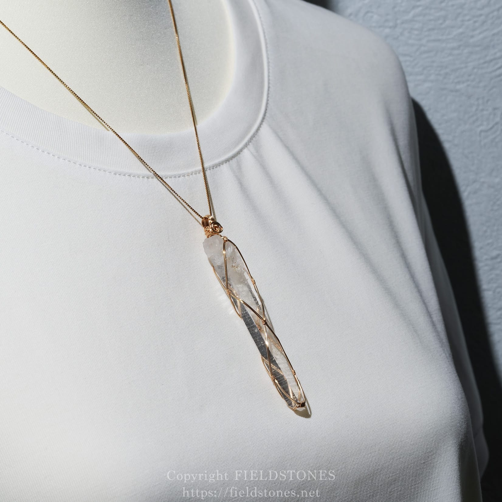 6751-8 - FieldStones Gold wire-wrapped crystal pendant necklace resting on a white fabric garment display.