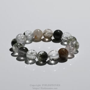 Clear gemstone beads with natural inclusions arranged in a circular bracelet on a reflective surface, creating mirrored shadows and highlights.