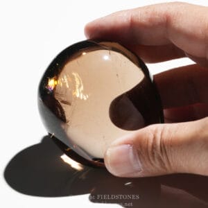Close-up of a hand holding a brown-tinted glass cabochon with scratches and reflections on a white background.