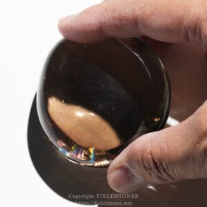 Close-up of a hand grasping a polished dark brown glass orb with subtle rainbow reflections along its edge.
