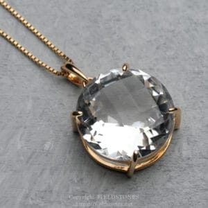 Gold pendant necklace featuring a large, round, faceted clear gemstone in a four-prong gold setting on a thin gold chain, resting on a gray surface.