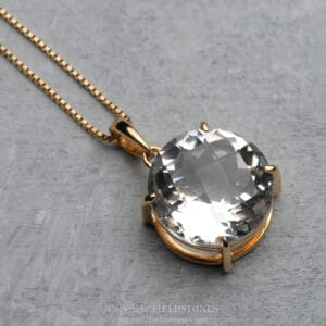 Gold-toned pendant necklace with a large faceted colorless gemstone on a delicate chain, resting on a gray surface.